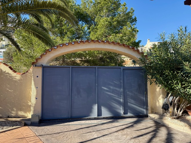 Puerta de entrada corredera metálica bajo un arco de estilo mediterráneo con tejas, rodeada de vegetación madura y suelo de hormigón impreso.