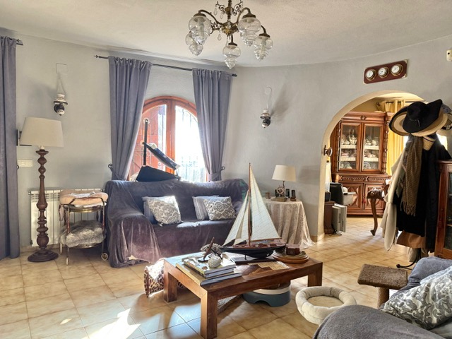 Salón con paredes curvas, suelos de gres y un arco que comunica con el comedor. Presenta una ventana de madera arqueada, lámpara de araña y calefacción por radiador.
