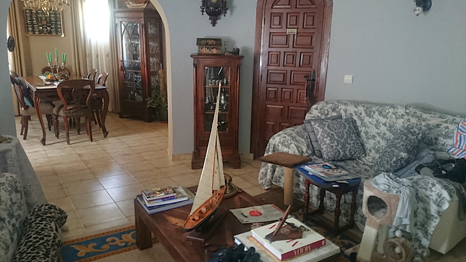 Salón tradicional con suelos cerámicos, arco hacia el comedor y mobiliario de madera clásica. Incluye vitrinas ornamentadas y una mesa de centro con detalles náuticos.