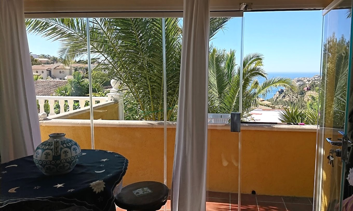 Terraza acristalada con cortinas de cristal, mobiliario auxiliar y vistas despejadas al mar Mediterráneo entre palmeras y vegetación autóctona.