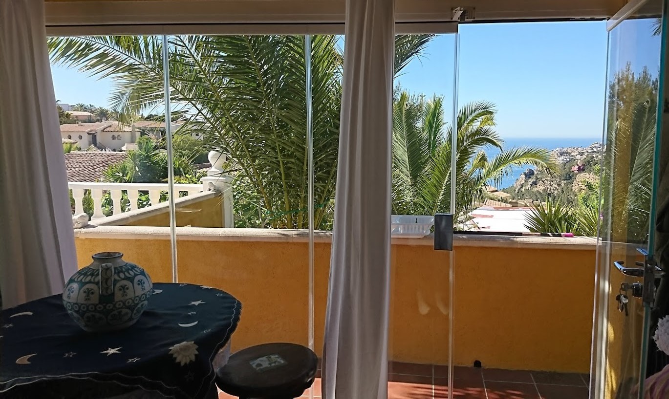 Terraza acristalada con cortinas de cristal que ofrece vistas al mar y palmeras. Incluye suelos de terracota y acceso directo al exterior.