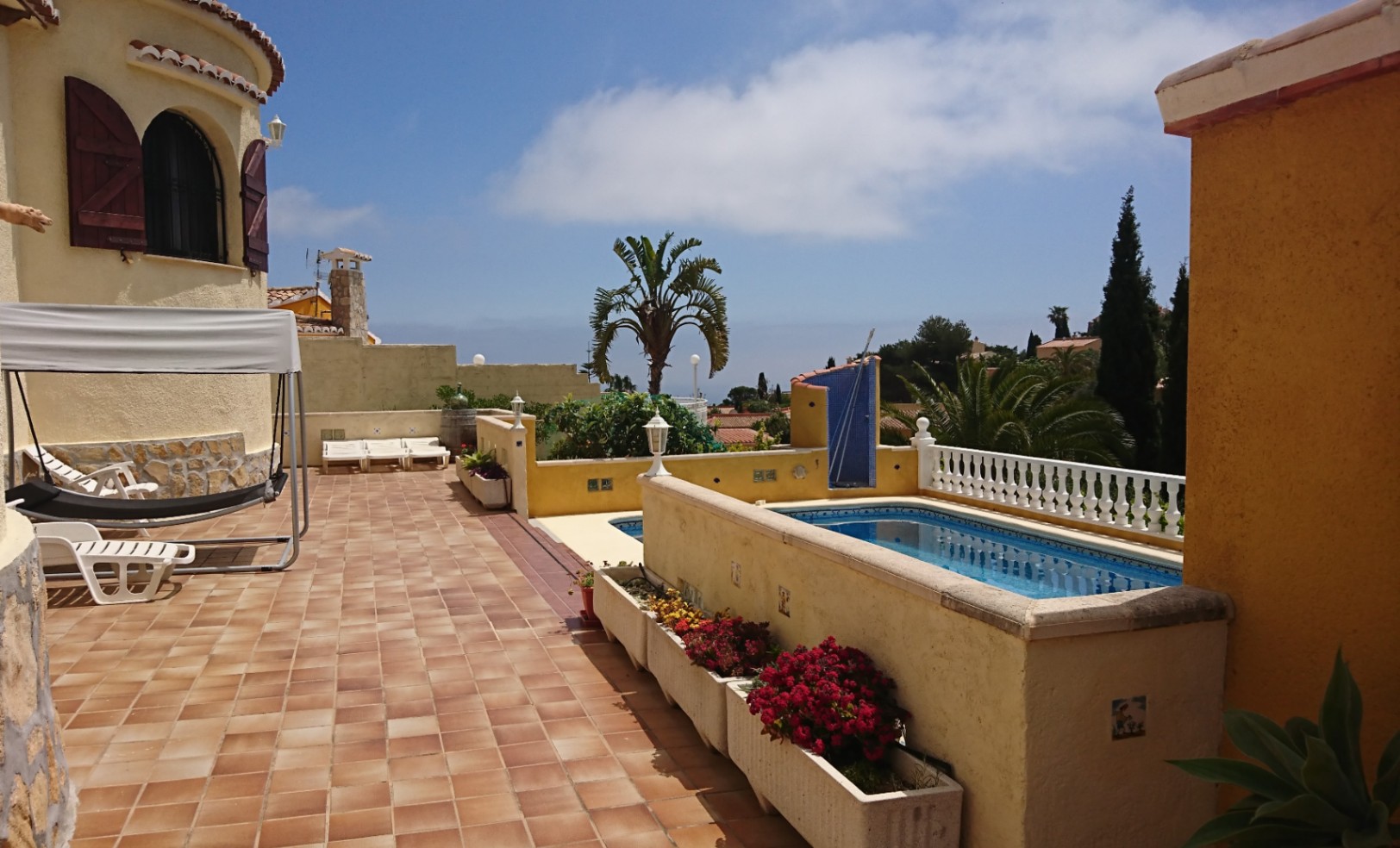 Terraza exterior con suelos de gres, piscina privada y zona de descanso. Destaca la torre de la villa con contraventanas de madera y vistas al entorno mediterráneo.