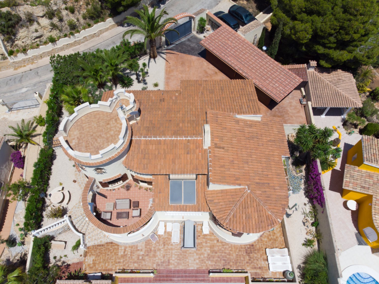 Vista aérea cenital de la villa mostrando su arquitectura mediterránea, la torre distintiva, tejados de terracota, paneles solares y amplias terrazas con mobiliario exterior.