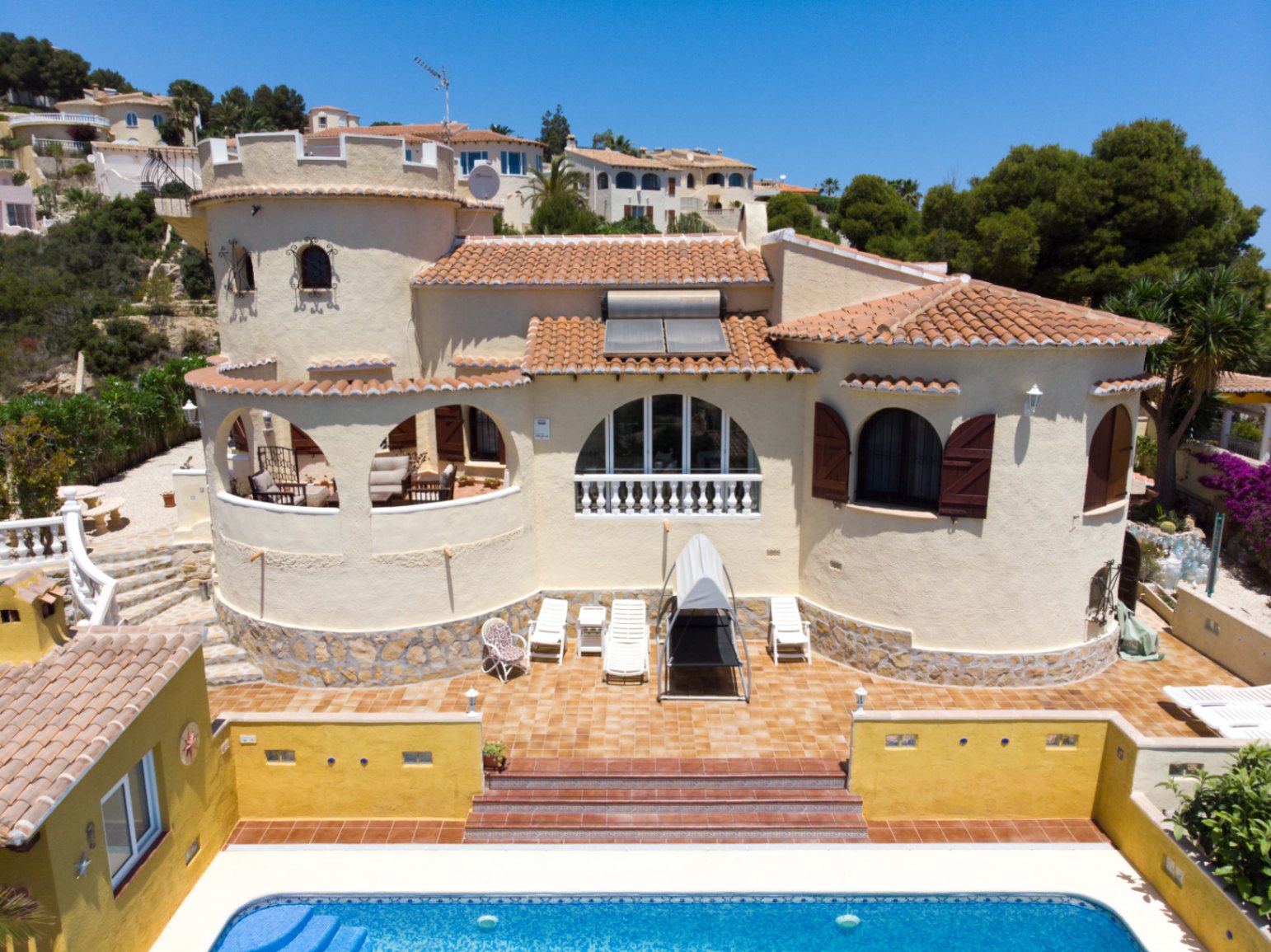 Vista exterior de la villa con torre circular, terraza de terracota y piscina. Destacan los arcos mediterráneos, persianas de madera y paneles solares sobre el tejado de teja.