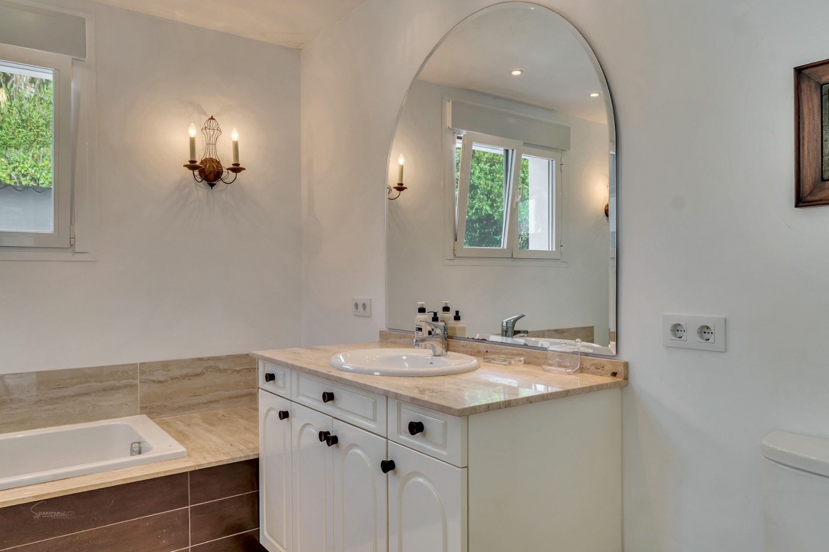 Baño con encimera de mármol, gran espejo de arco y apliques clásicos. Incluye bañera integrada y ventana con vistas al jardín, creando un ambiente luminoso y funcional.