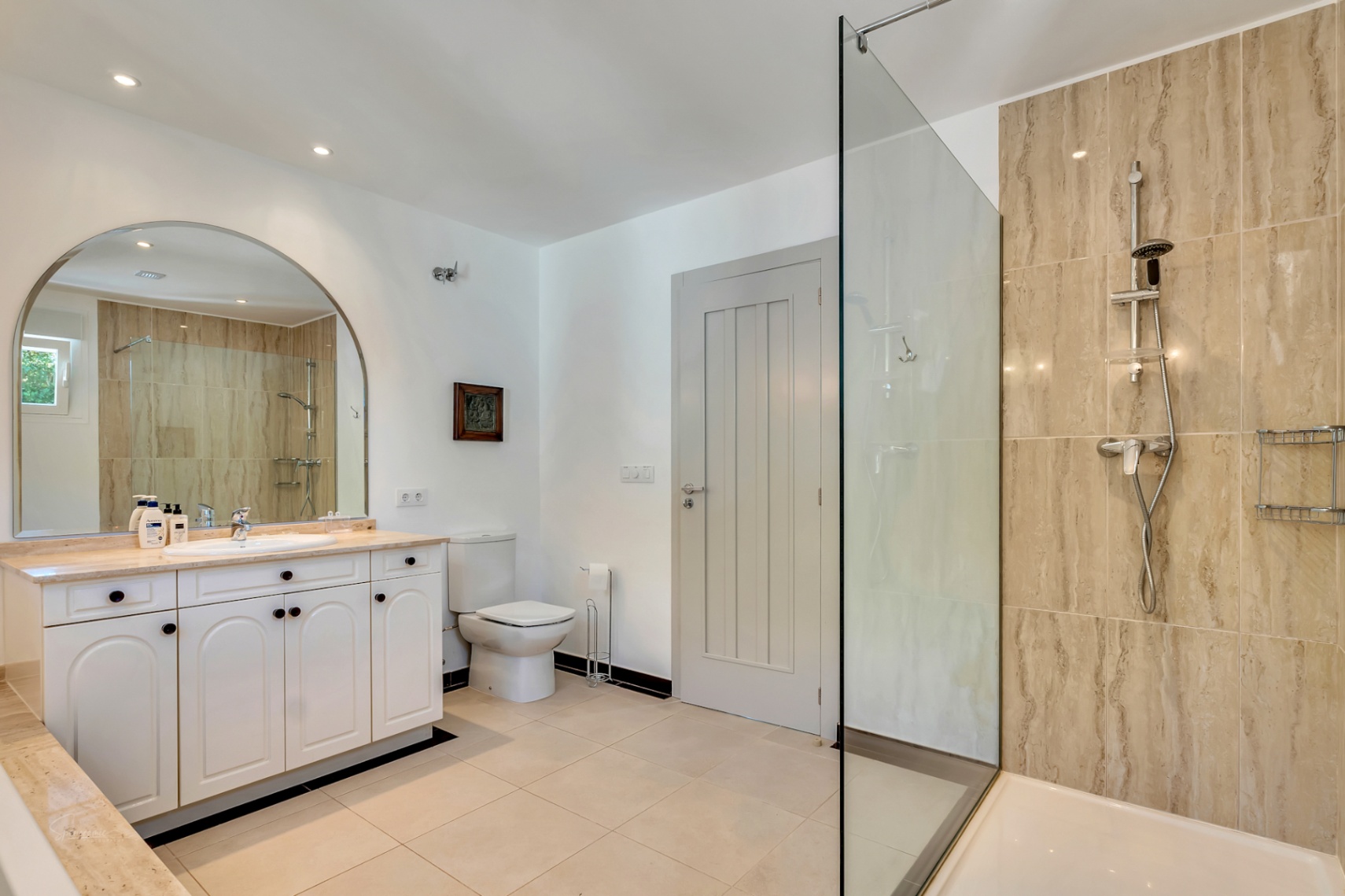 Baño elegante con ducha de obra revestida en mármol beige, mampara de cristal, lavabo con encimera de piedra y un gran espejo de arco que amplía el espacio.