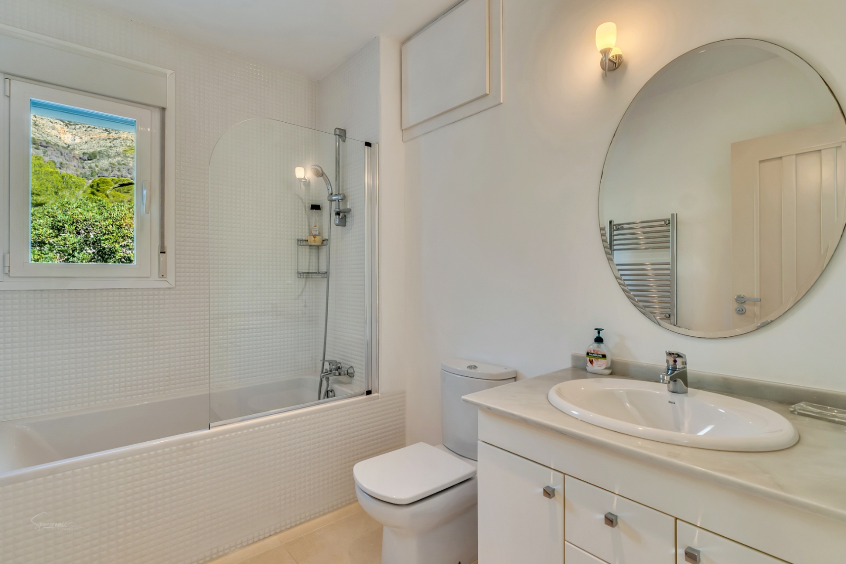Baño luminoso con azulejos de mosaico blanco, encimera de mármol y bañera con mampara de cristal. La ventana ofrece vistas directas a la montaña Montgó y la vegetación circundante.