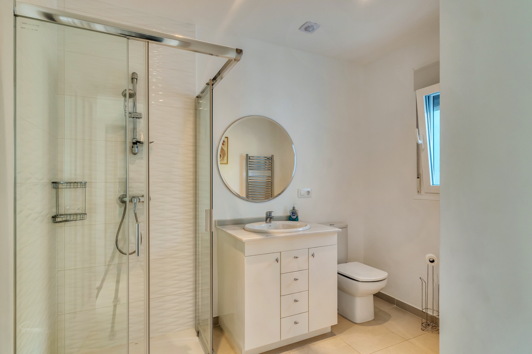 Baño moderno con ducha de obra acristalada, azulejos blancos con relieve, lavabo con mueble blanco, espejo circular y luz natural a través de una ventana lateral.