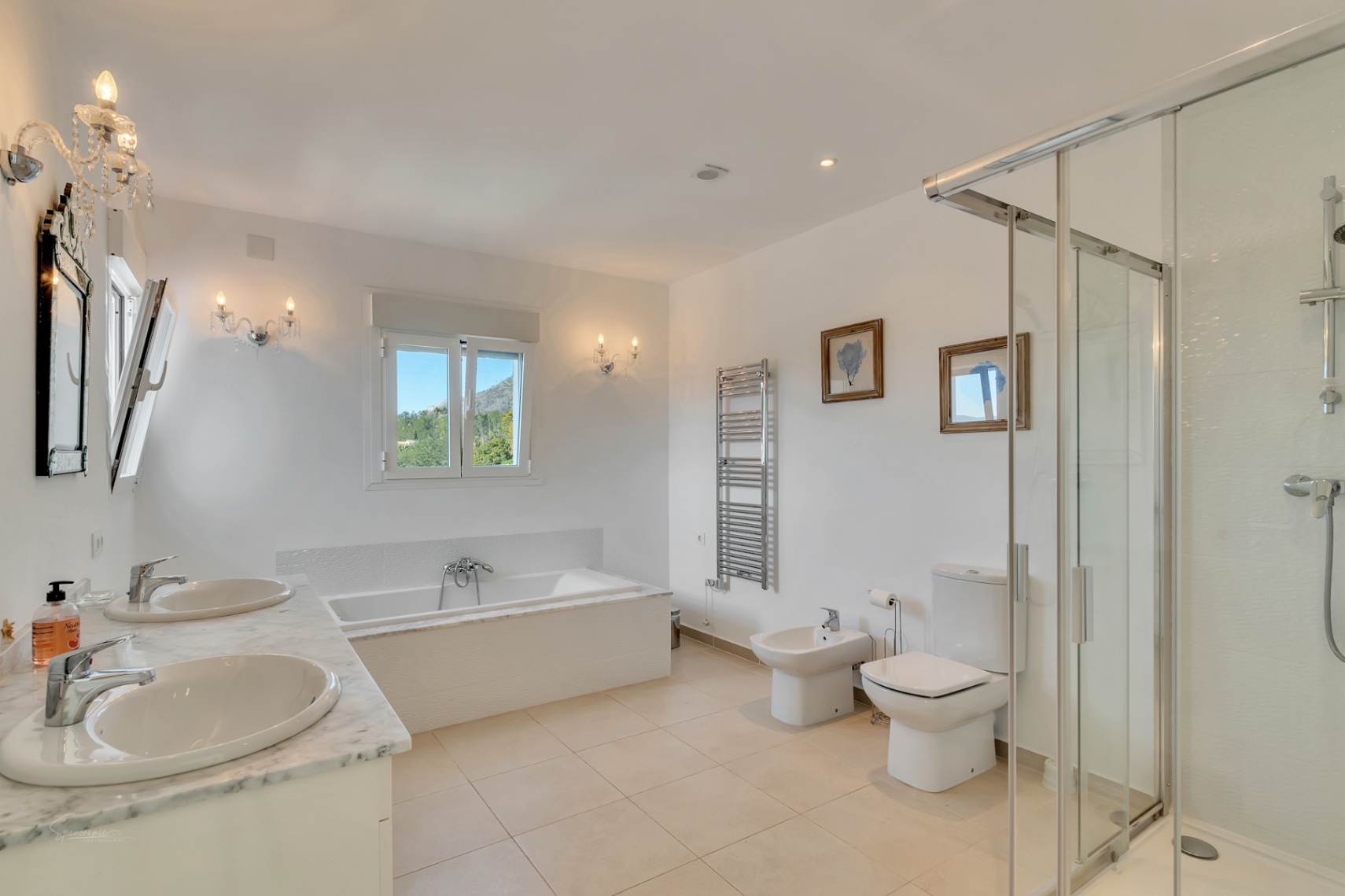 Baño principal con encimera de mármol de doble seno, bañera encastrada, ducha independiente y elegantes apliques de cristal con vistas al Montgó.