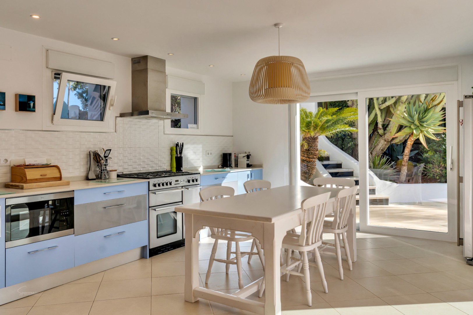 Cocina equipada con armarios azul pastel, encimera blanca y mesa de madera, conectada al exterior mediante amplios ventanales que iluminan el espacio.