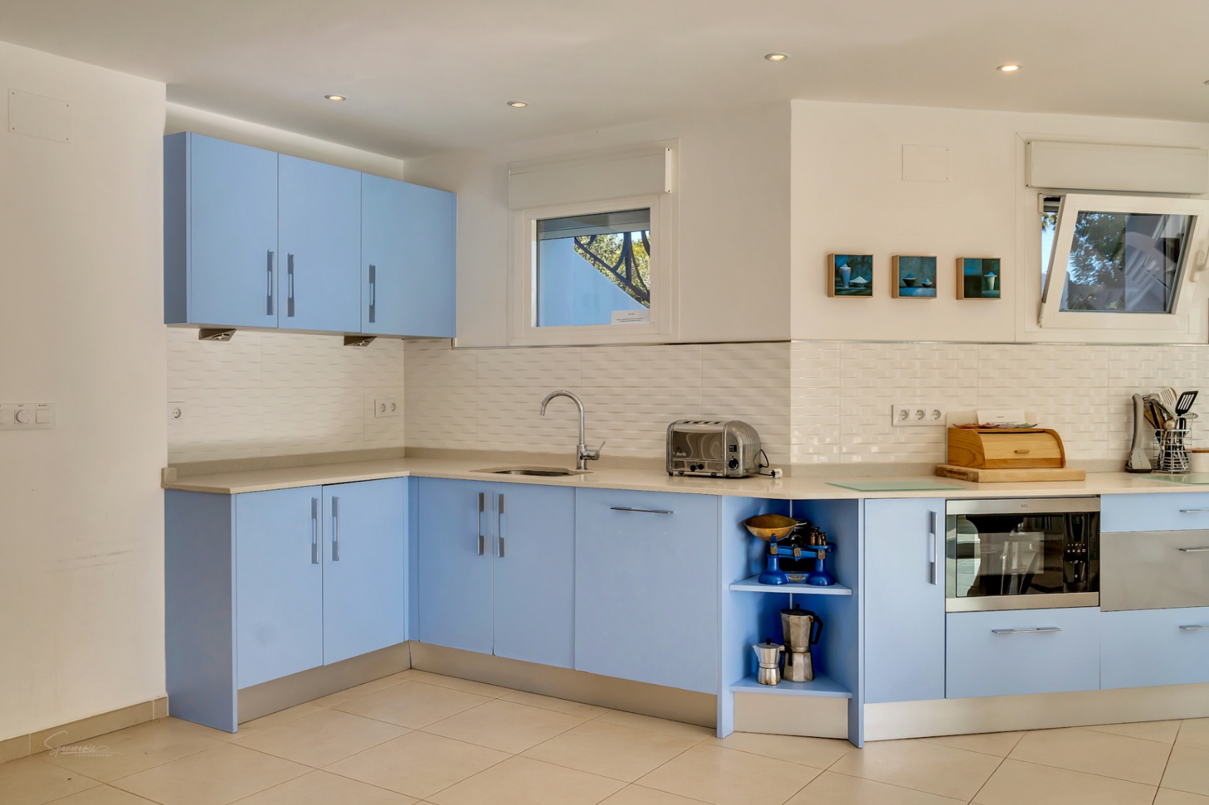 Cocina funcional con armarios azul pastel, encimera de piedra clara y salpicadero de azulejos blancos. Dispone de electrodomésticos integrados y ventanas que aportan luz natural.