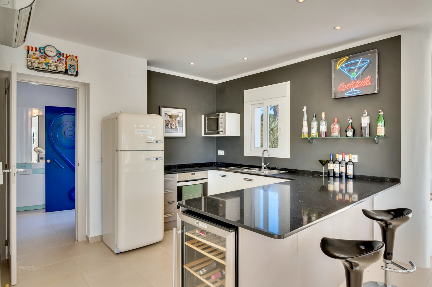 Cocina moderna con encimera de granito negro, nevera Smeg de estilo retro y zona de bar con vinoteca integrada y cartel de neón decorativo.
