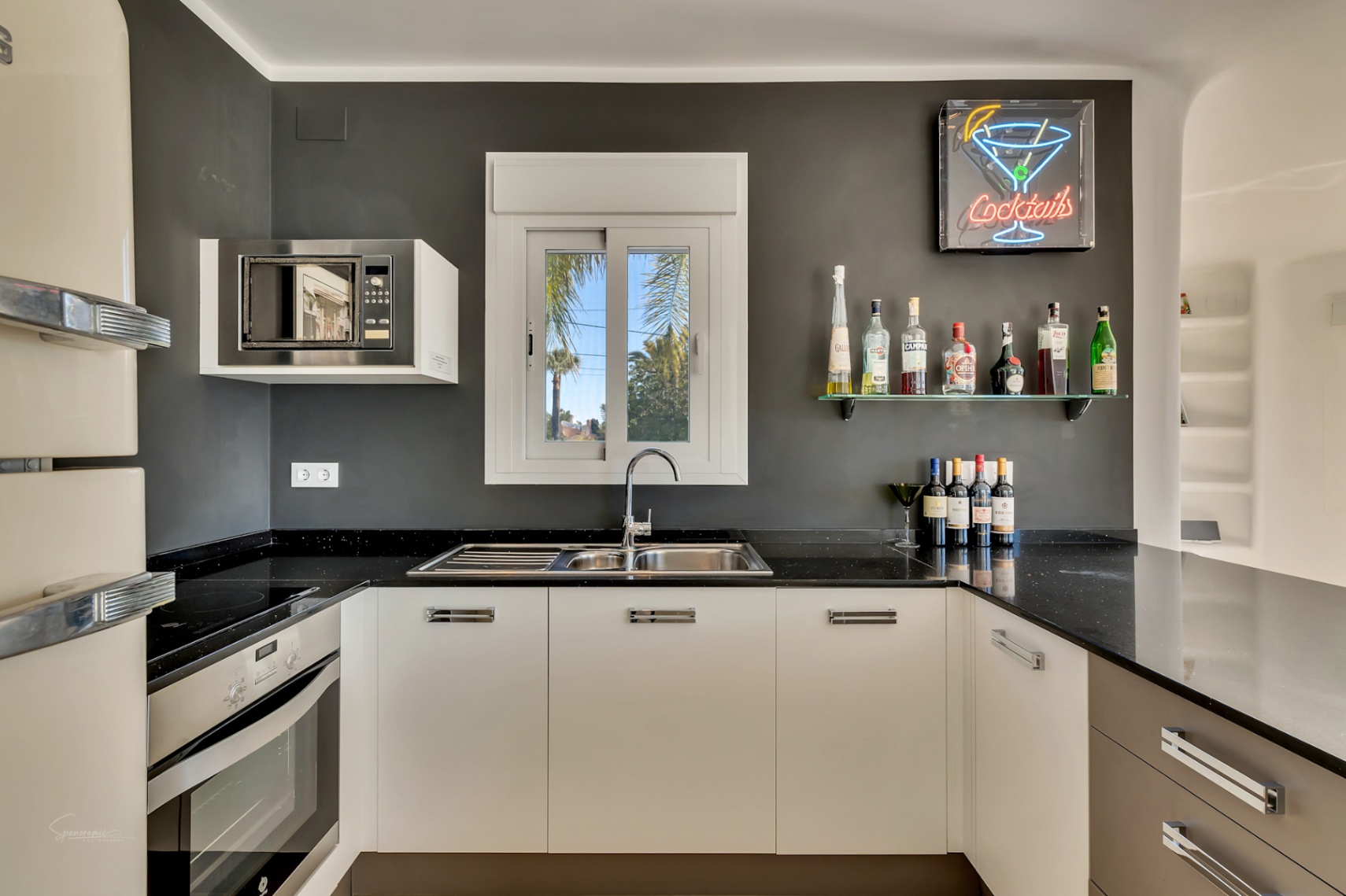 Cocina moderna con encimera negra, armarios blancos y zona de bar con estante de cristal y letrero de neón. Incluye electrodomésticos integrados y vistas al jardín.