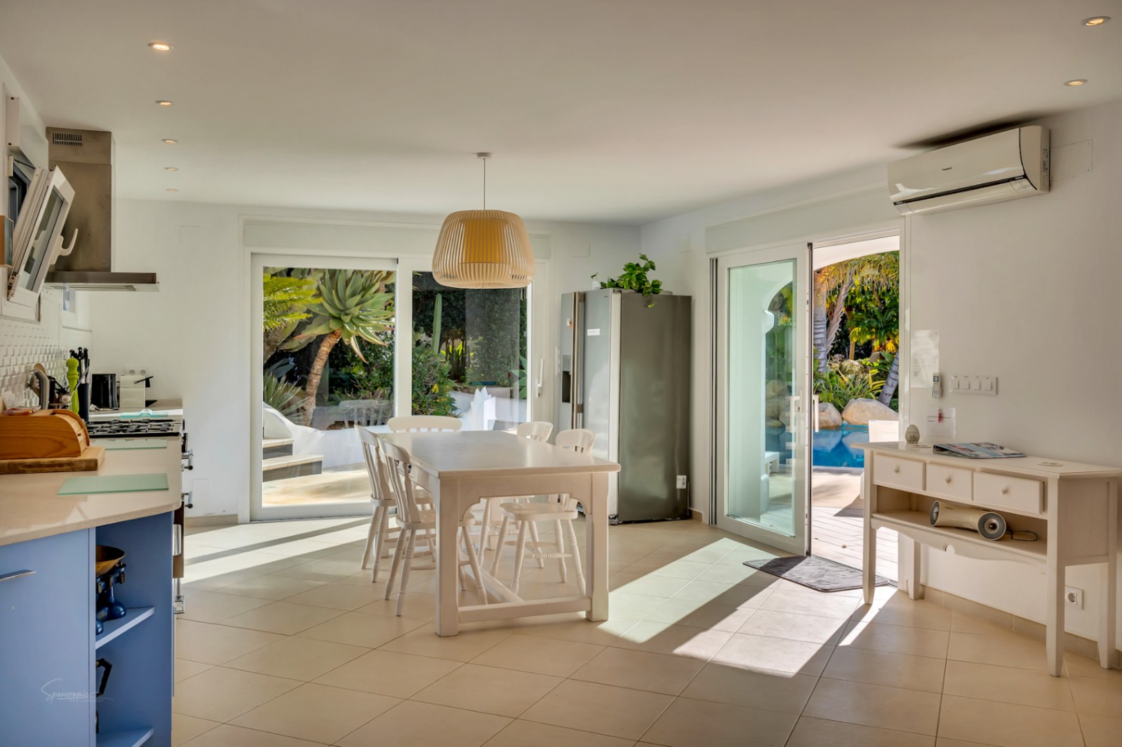 Cocina y comedor de planta abierta con muebles blancos y detalles en azul. Las puertas correderas de cristal conectan el interior con la piscina y el jardín mediterráneo.