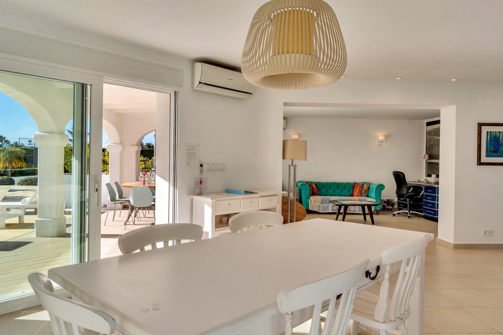 Comedor luminoso con mesa de madera blanca y lámpara de mimbre, conectado a una zona de estar con sofá turquesa, despacho integrado y acceso directo a la naya.