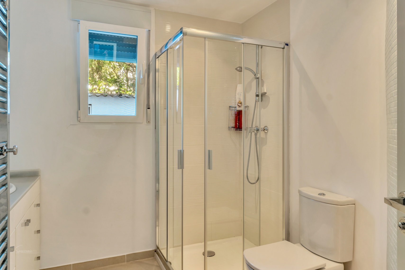 Cuarto de baño moderno con cabina de ducha acristalada, mueble lavabo blanco con cajones y ventana que aporta luz natural y vistas al exterior.
