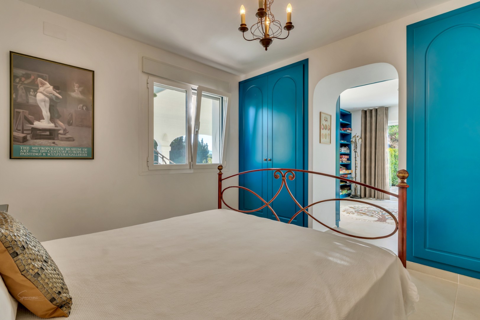 Dormitorio con armarios empotrados en azul vibrante, cama de forja y acceso directo a un estudio privado a través de un elegante arco arquitectónico.