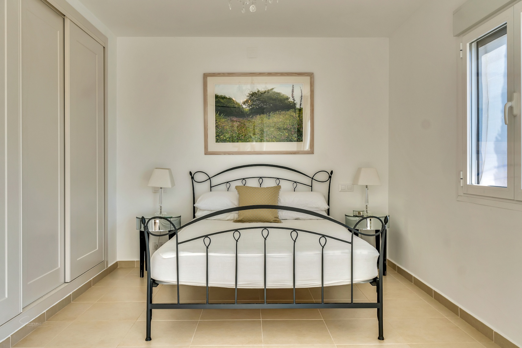 Dormitorio con cama de hierro forjado negro, armarios empotrados en tono crema y suelos cerámicos. Dispone de mesitas de noche de espejo y luz natural lateral.