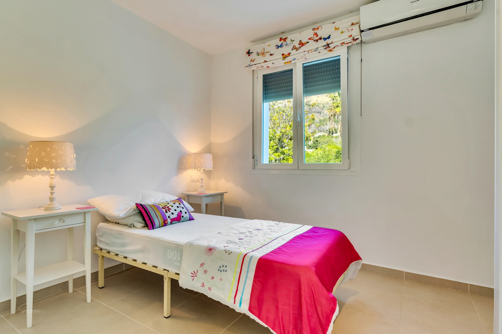 Dormitorio con cama individual, aire acondicionado split y ventana con vistas al jardín. Incluye mesitas de noche blancas y lámparas con detalles de mariposas.