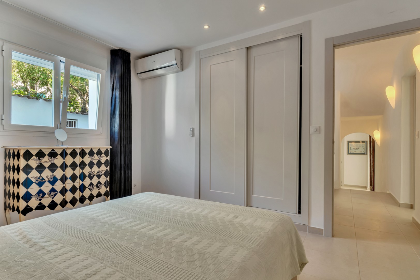 Dormitorio luminoso con armarios empotrados, aire acondicionado y una cómoda decorativa. La estancia conecta con un pasillo iluminado por apliques de diseño.