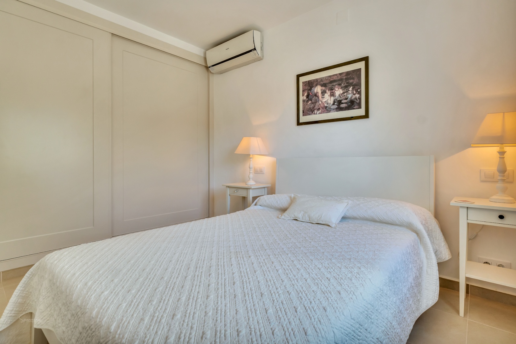 Dormitorio luminoso con armarios empotrados de puertas correderas, aire acondicionado individual y mesitas de noche bajo una iluminación cálida.