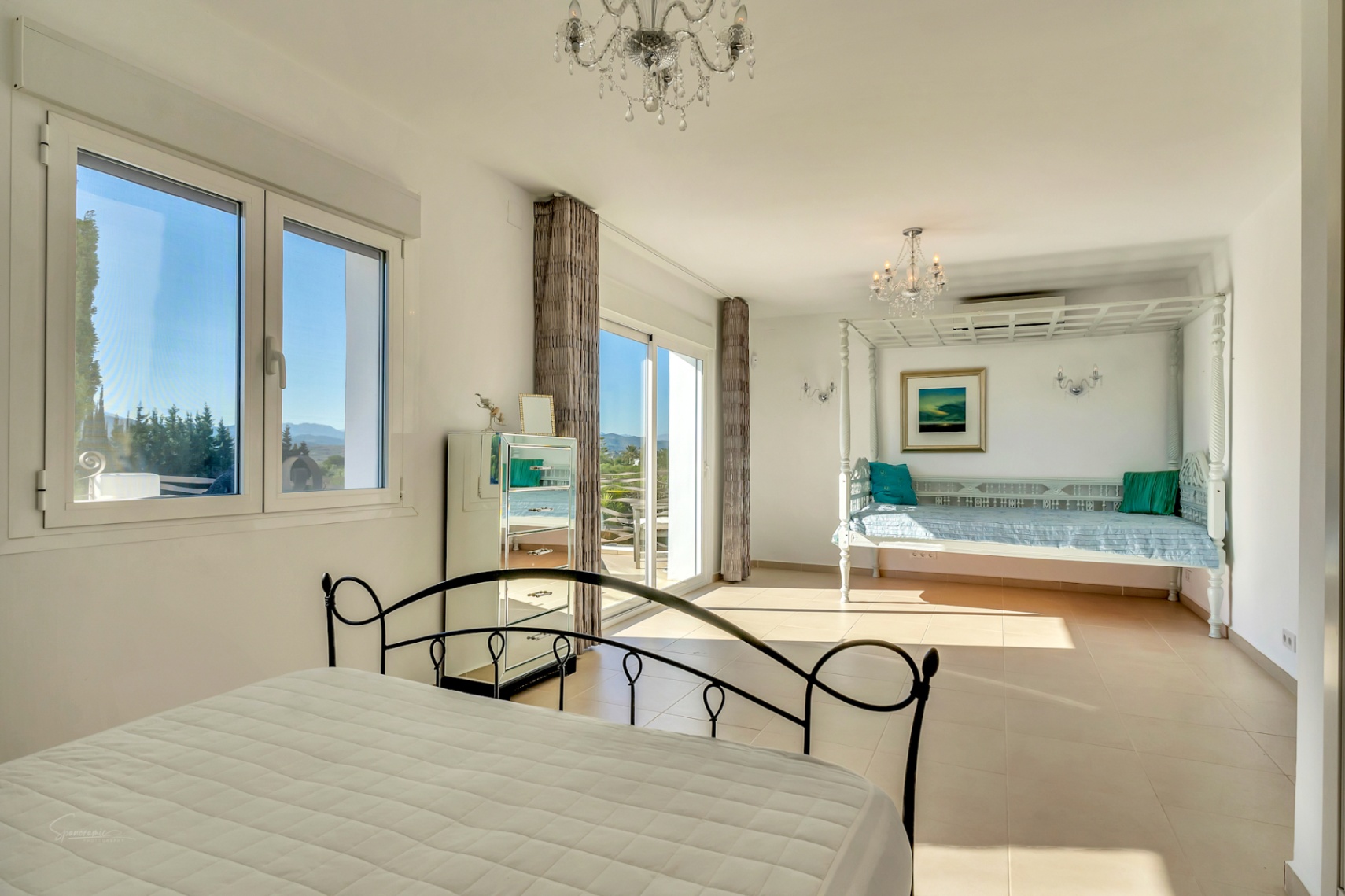 Dormitorio luminoso con cama de forja, cómoda de espejo y un diván de madera tallada bajo lámparas de cristal, con acceso directo a la terraza exterior.