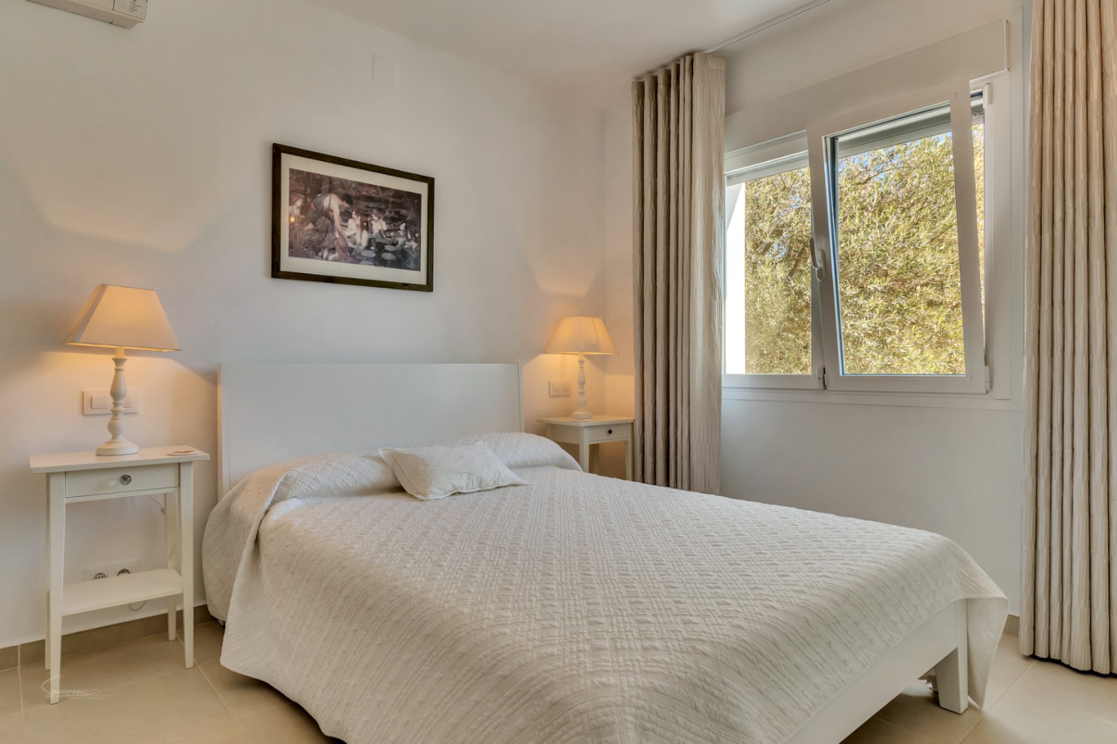 Dormitorio luminoso con cama doble blanca, mesitas de noche a juego y ventana con vistas al jardín. Incluye iluminación cálida y textiles en tonos claros.