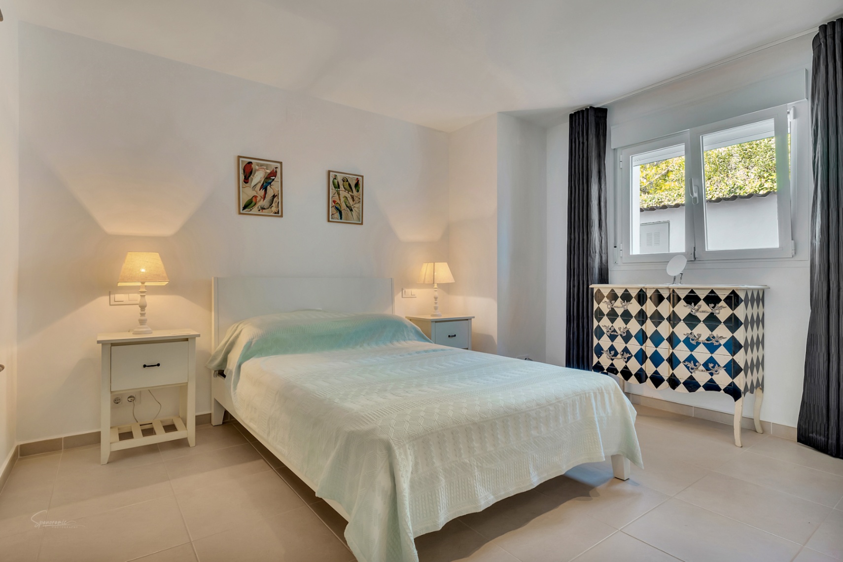 Dormitorio luminoso con cama doble, mesitas de noche blancas y una cómoda de diseño con patrón de rombos. Incluye suelos cerámicos claros, luz natural y aire acondicionado.