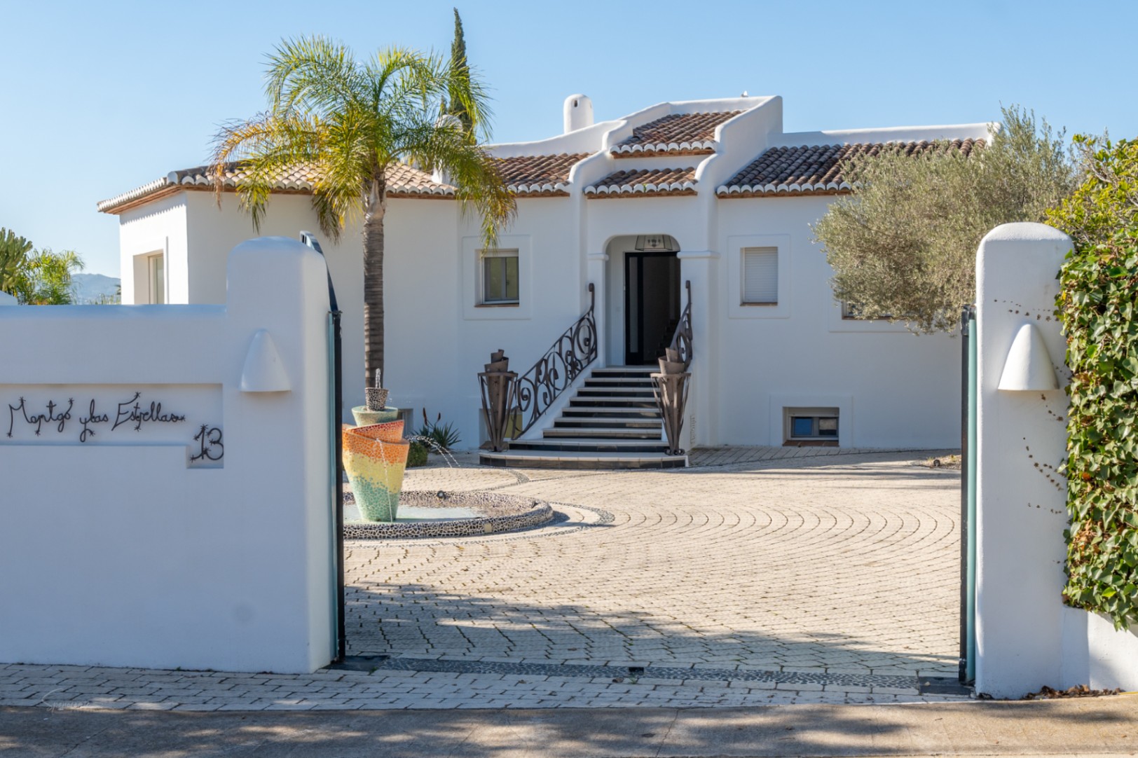 Entrada de estilo ibicenco con fachada blanca, fuente central de mosaico y patio pavimentado. Destacan las barandillas de hierro forjado y la arquitectura de líneas redondeadas.