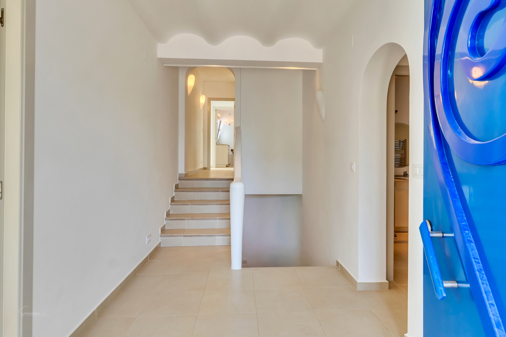Entrada principal con una puerta azul de diseño circular, techos con molduras onduladas y acceso a los dos niveles de la villa mediante escaleras.