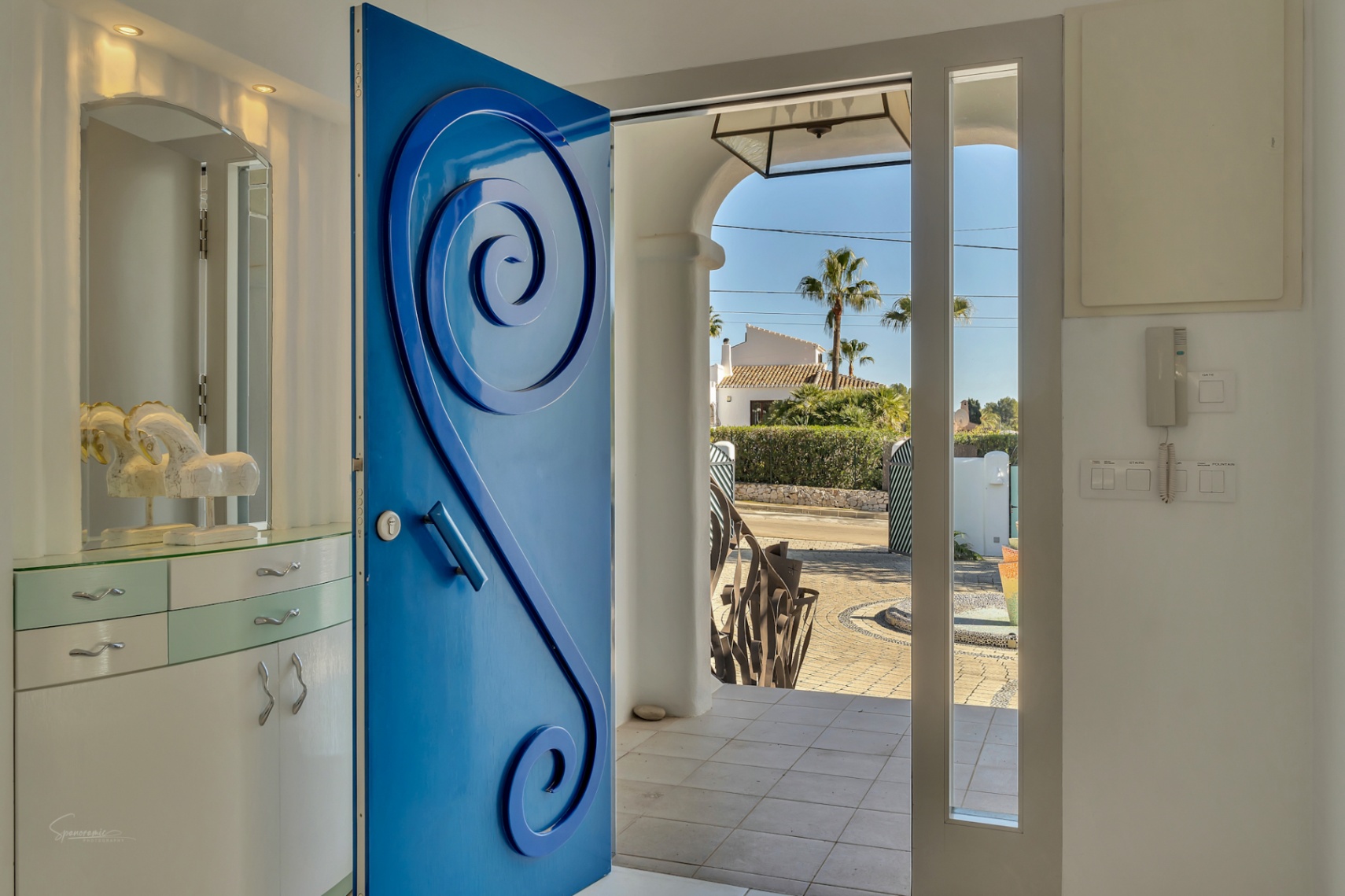 Entrada principal con una puerta azul de diseño en espiral que da paso al patio pavimentado y jardines exteriores bajo el sol mediterráneo.