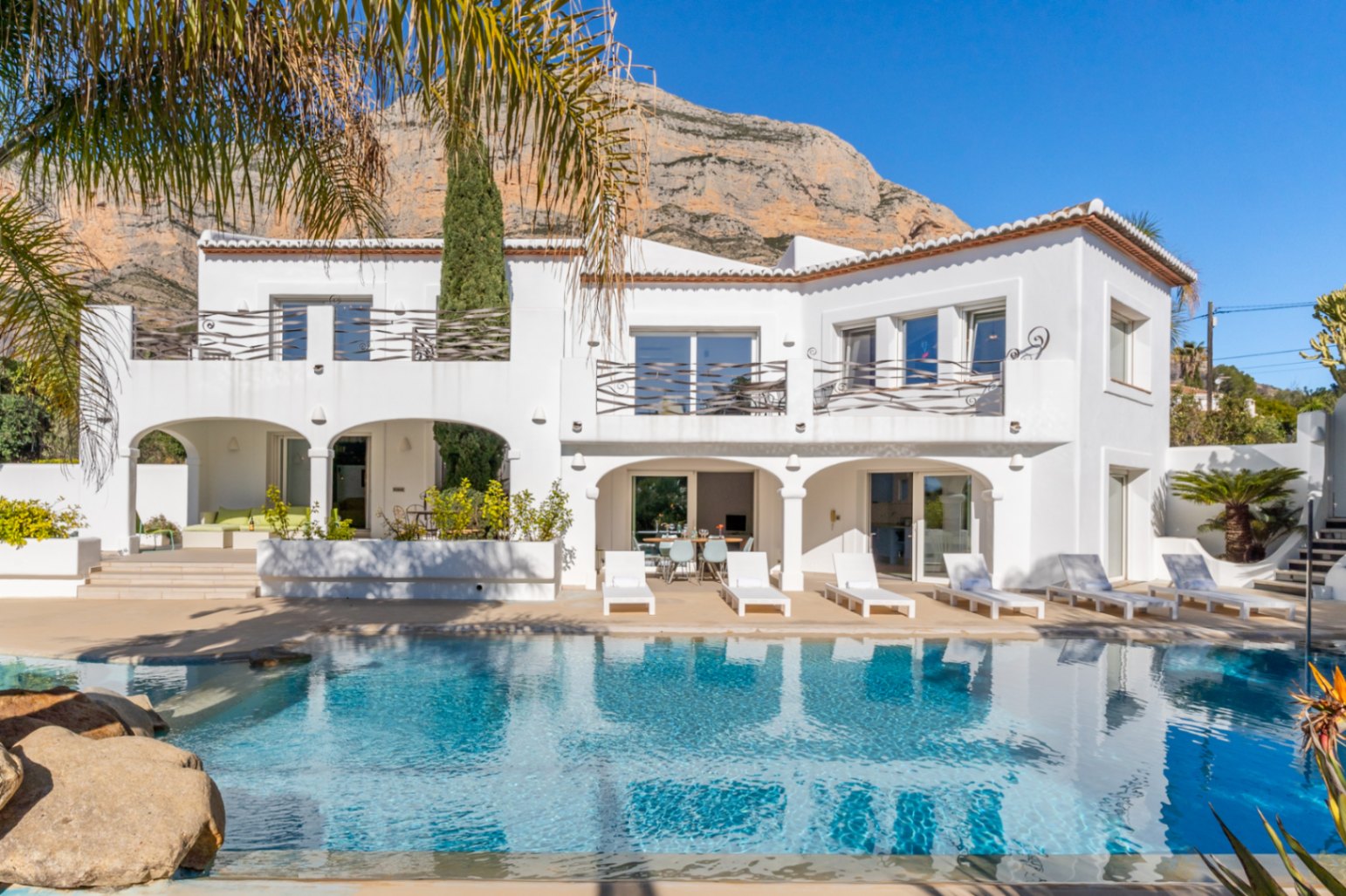Fachada blanca de estilo ibicenco con piscina, naya de arcos y terrazas superiores bajo la imponente silueta de la montaña Montgó.