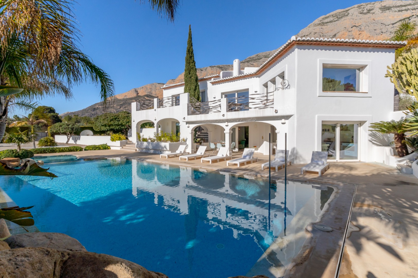 Fachada posterior de la villa estilo Ibiza con piscina de 70m², naya de arcos y terraza superior con barandillas de hierro frente al Montgó.