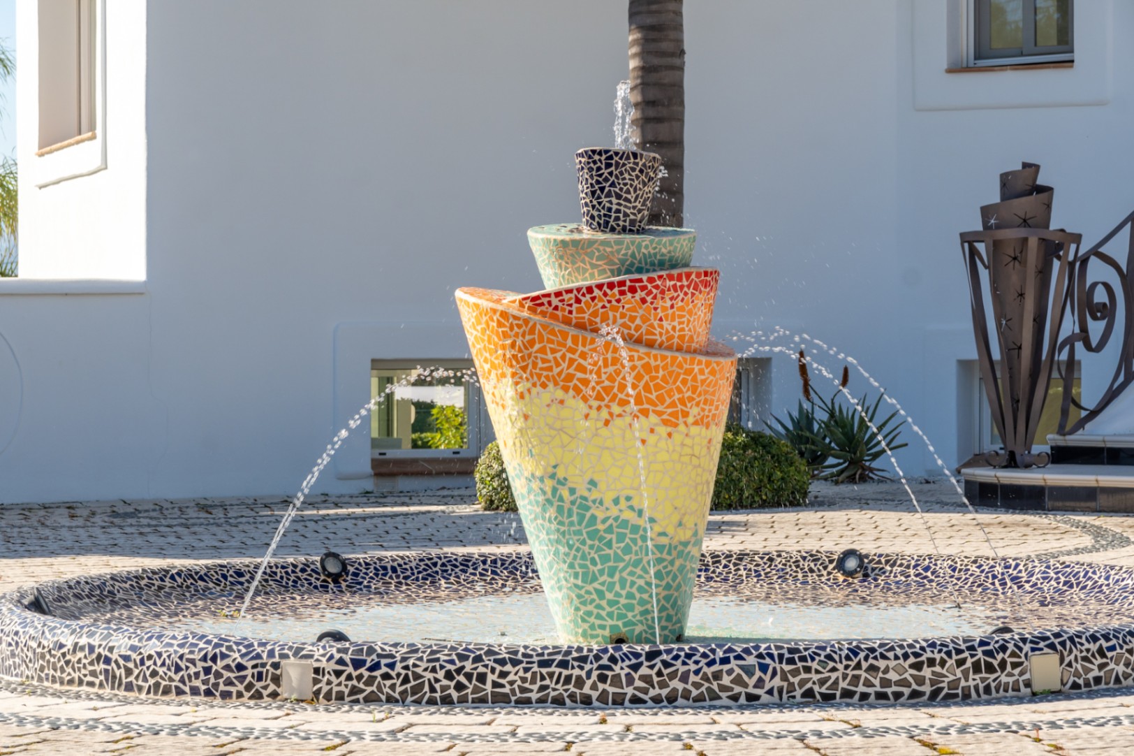 Fuente central de diseño contemporáneo con mosaicos multicolores, situada en un patio pavimentado frente a la fachada blanca de la villa.