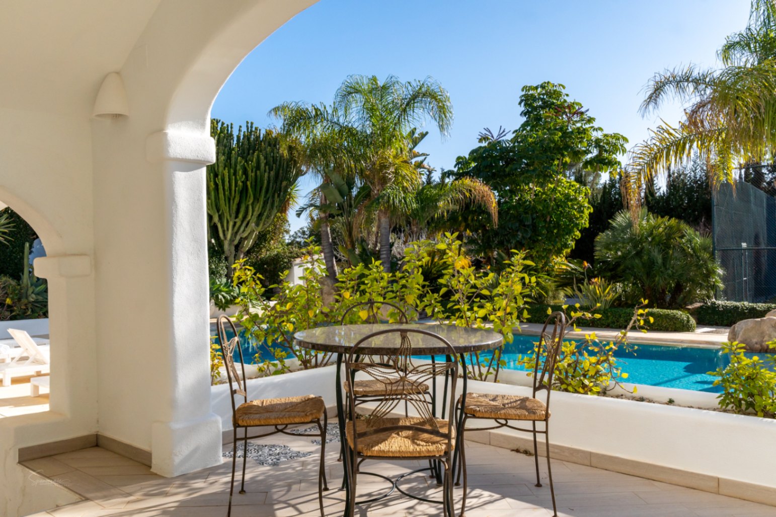 Naya cubierta con arcos blancos y mobiliario de hierro forjado, con vistas directas a la piscina y al jardín mediterráneo con palmeras y cactus.