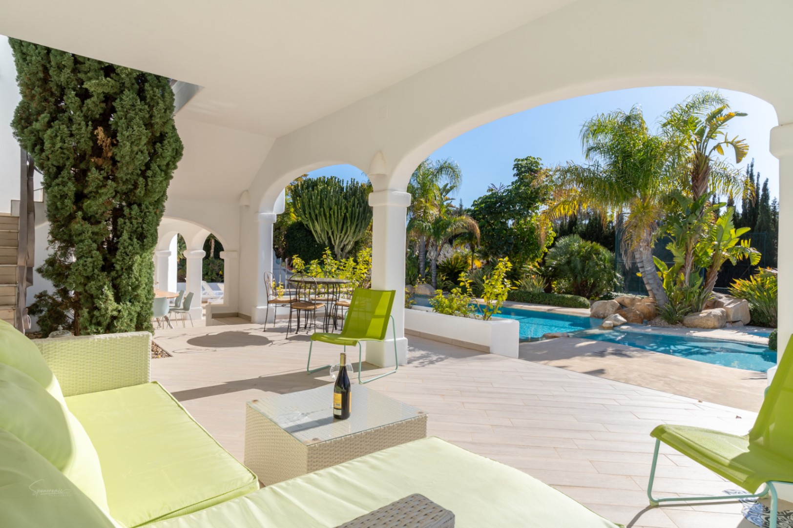 Naya cubierta con arcos blancos y suelos cerámicos, equipada con mobiliario de descanso frente a la piscina y el jardín mediterráneo con palmeras.
