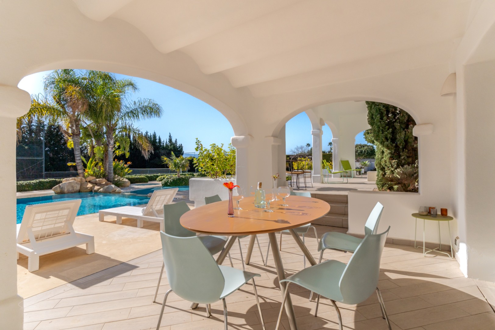 Naya cubierta con arcos tradicionales y comedor exterior, con vistas a la piscina con elementos de roca, palmeras y la pista de tenis privada.