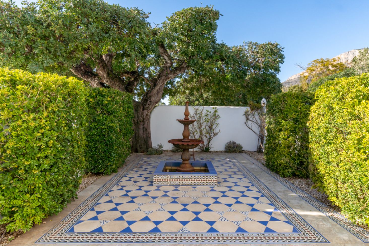 Patio exterior con fuente de estilo mediterráneo y suelo de baldosas geométricas en azul y blanco, rodeado de setos frondosos y con vistas al Montgó.