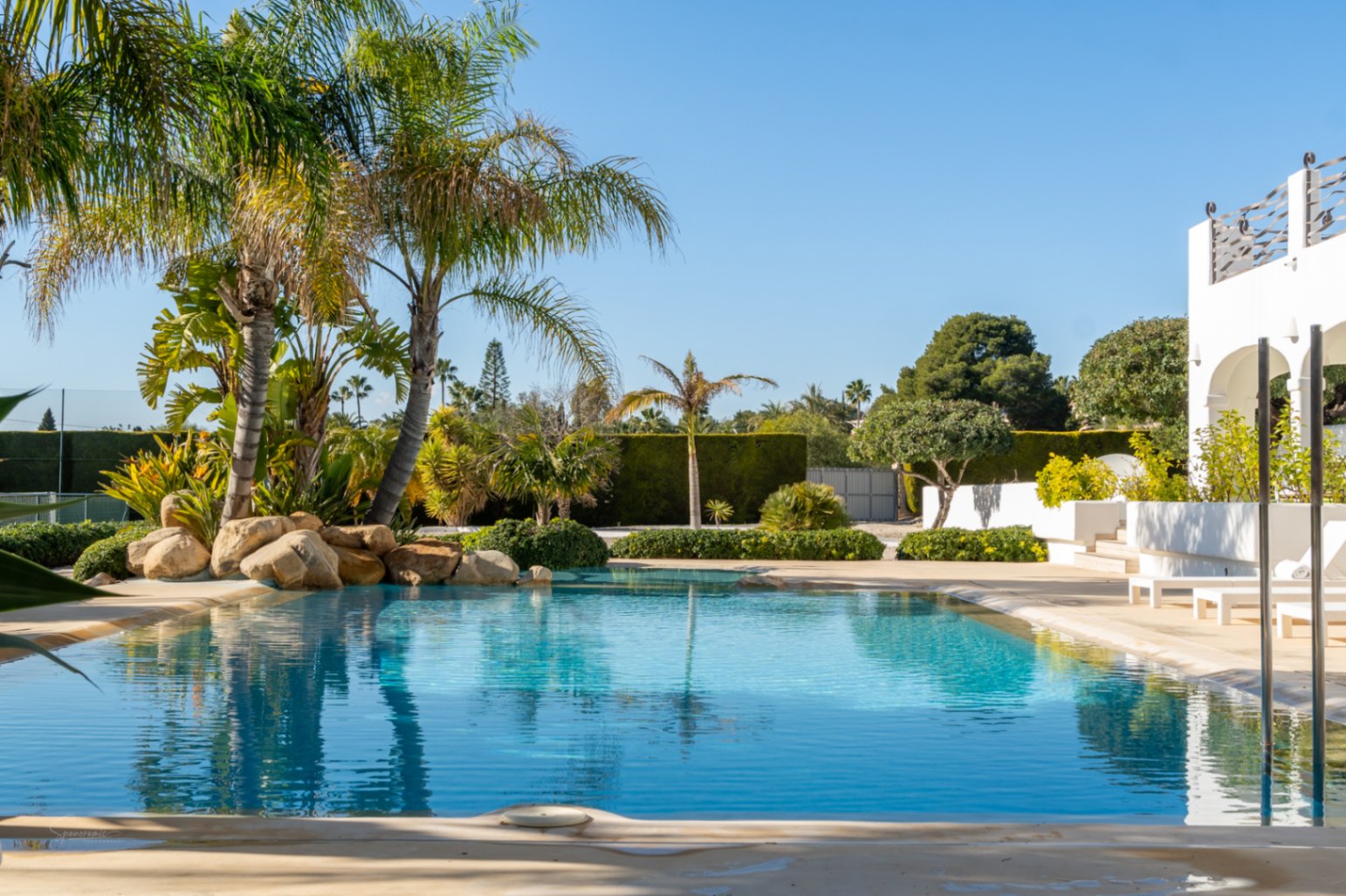 Piscina de diseño con entrada gradual, rodeada de palmeras y rocas naturales, junto a la naya arqueada de la villa y una pista de tenis privada.