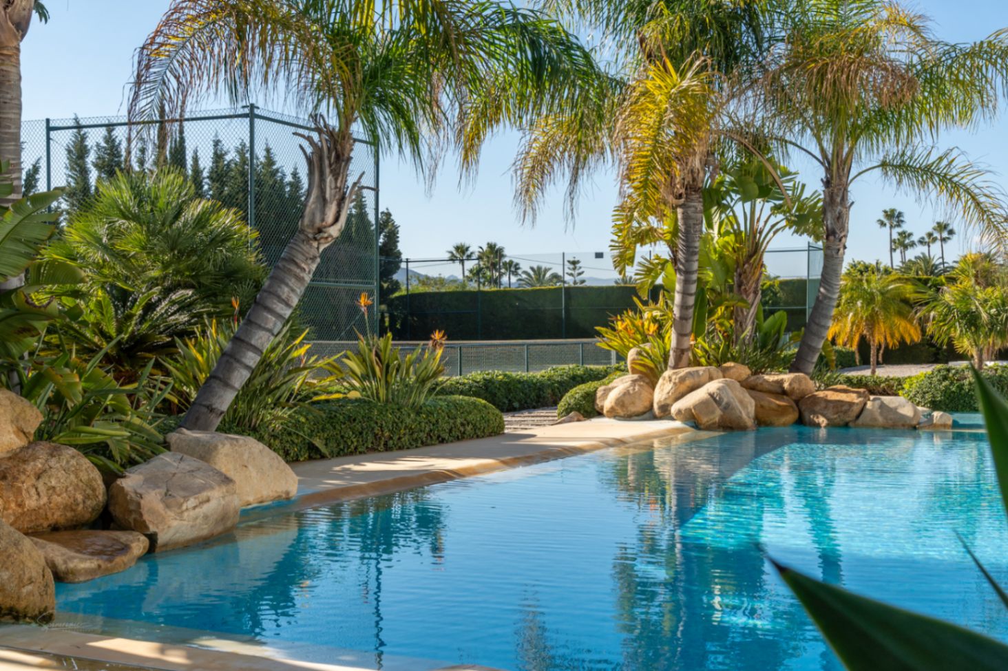 Piscina de diseño natural con bordes de roca y vegetación tropical, situada junto a la pista de tenis privada rodeada de palmeras.