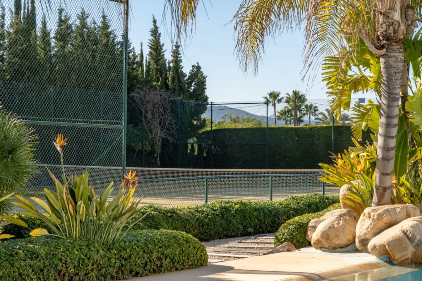 Pista de tenis privada rodeada de setos altos y vegetación mediterránea, con vistas al Montgó y un entorno ajardinado junto a la piscina.