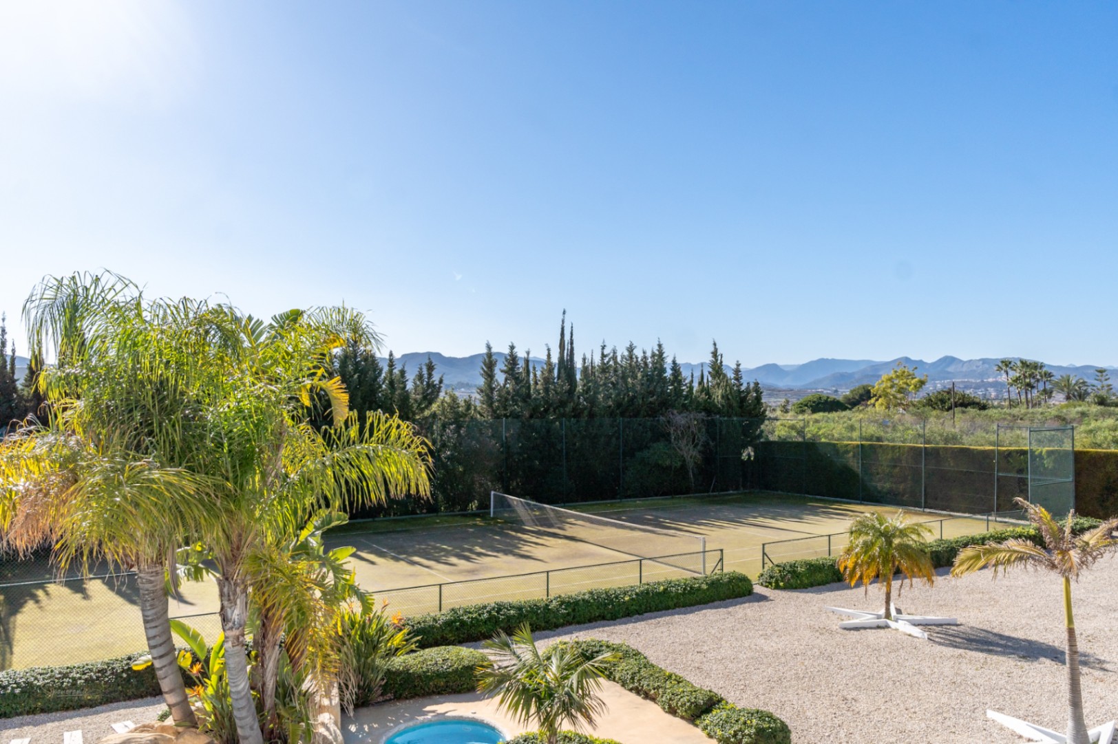 Pista de tenis privada rodeada de setos maduros y palmeras, con vistas panorámicas al valle y las montañas bajo un cielo despejado.