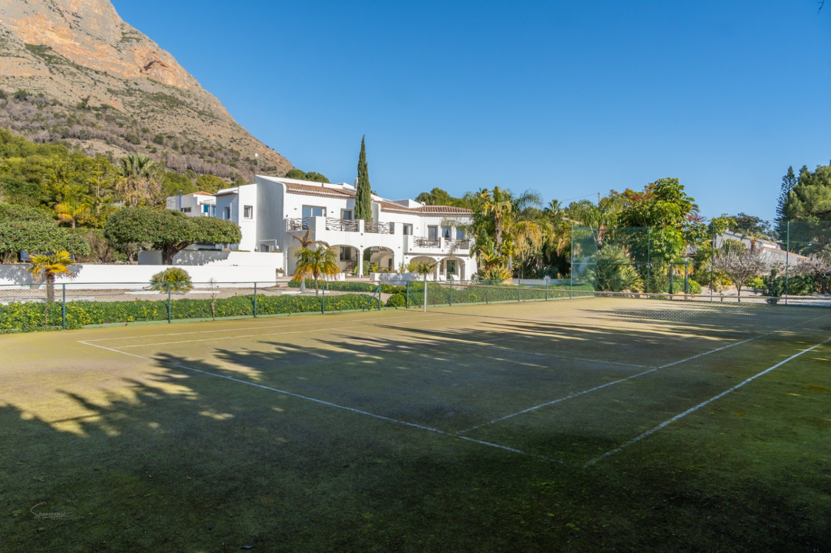 Pista de tenis privada rodeada de vegetación, con la villa de estilo ibicenco y el macizo del Montgó como telón de fondo bajo un cielo despejado.