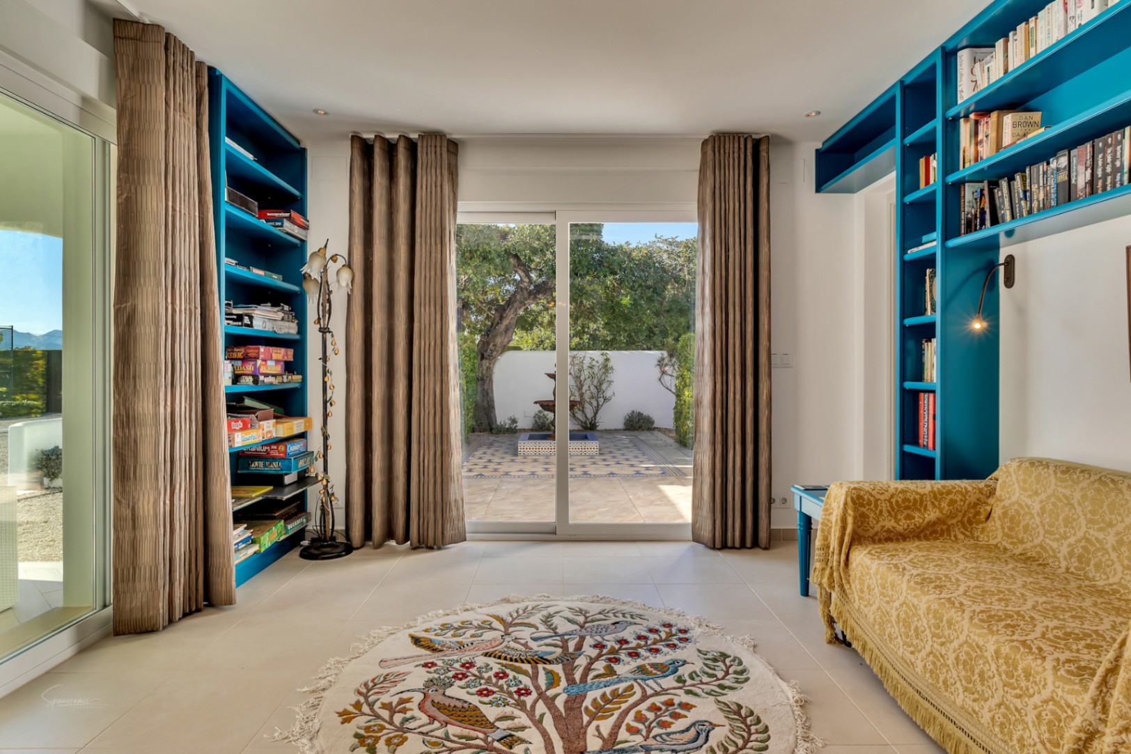 Sala de estar o despacho con estanterías integradas en azul vibrante, acceso directo a un patio con fuente y una alfombra decorativa con motivos naturales.
