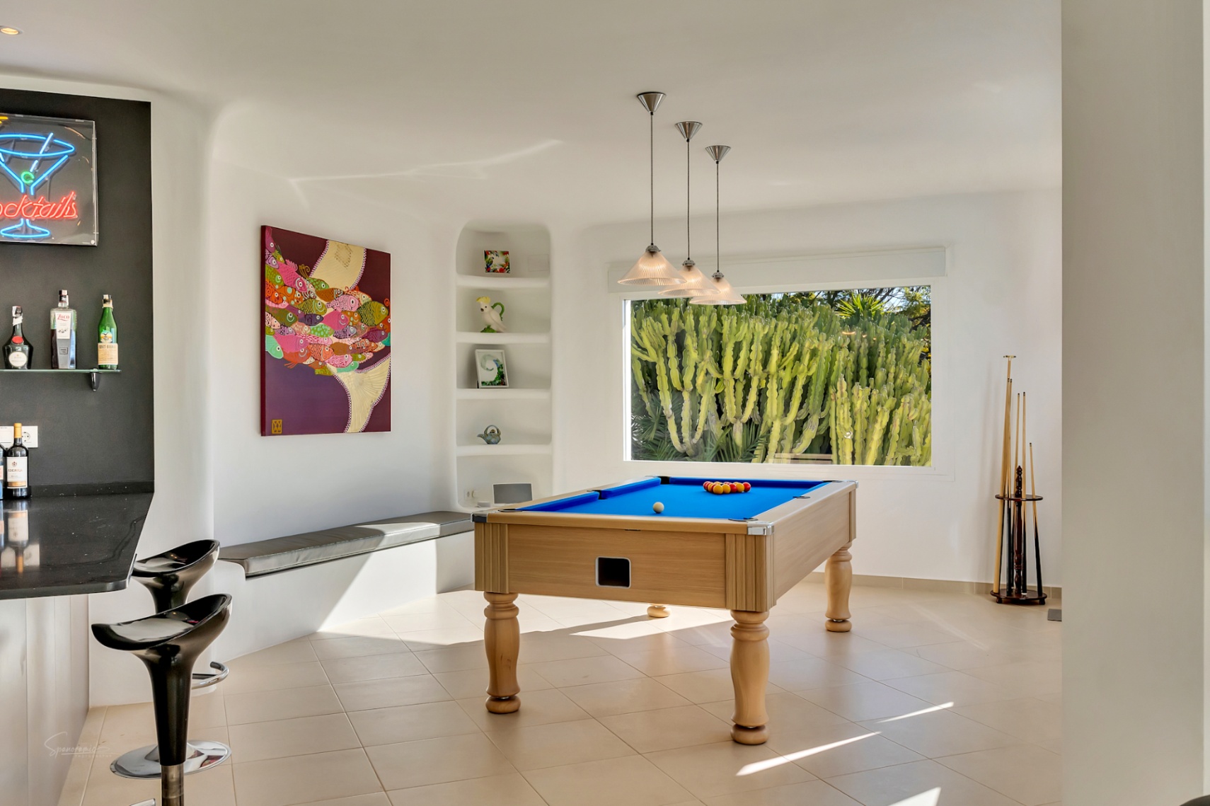 Sala de juegos de estilo ibicenco con mesa de billar, zona de bar integrada y ventanal con vistas a un jardín de cactus. Presenta paredes blancas redondeadas y abundante luz natural.