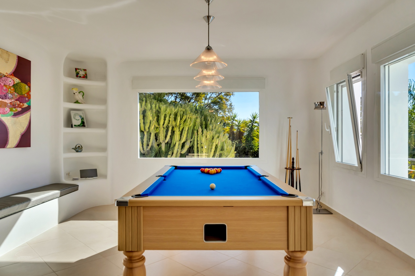 Sala de juegos luminosa con mesa de billar de madera y tapete azul, estanterías integradas y ventanal con vistas a un jardín de cactus.