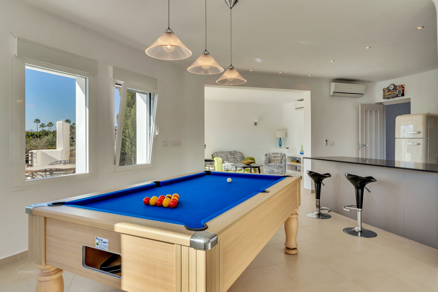 Sala de ocio con mesa de billar de tapete azul, barra con taburetes y nevera de diseño retro, integrada en un espacio luminoso con vistas al exterior.