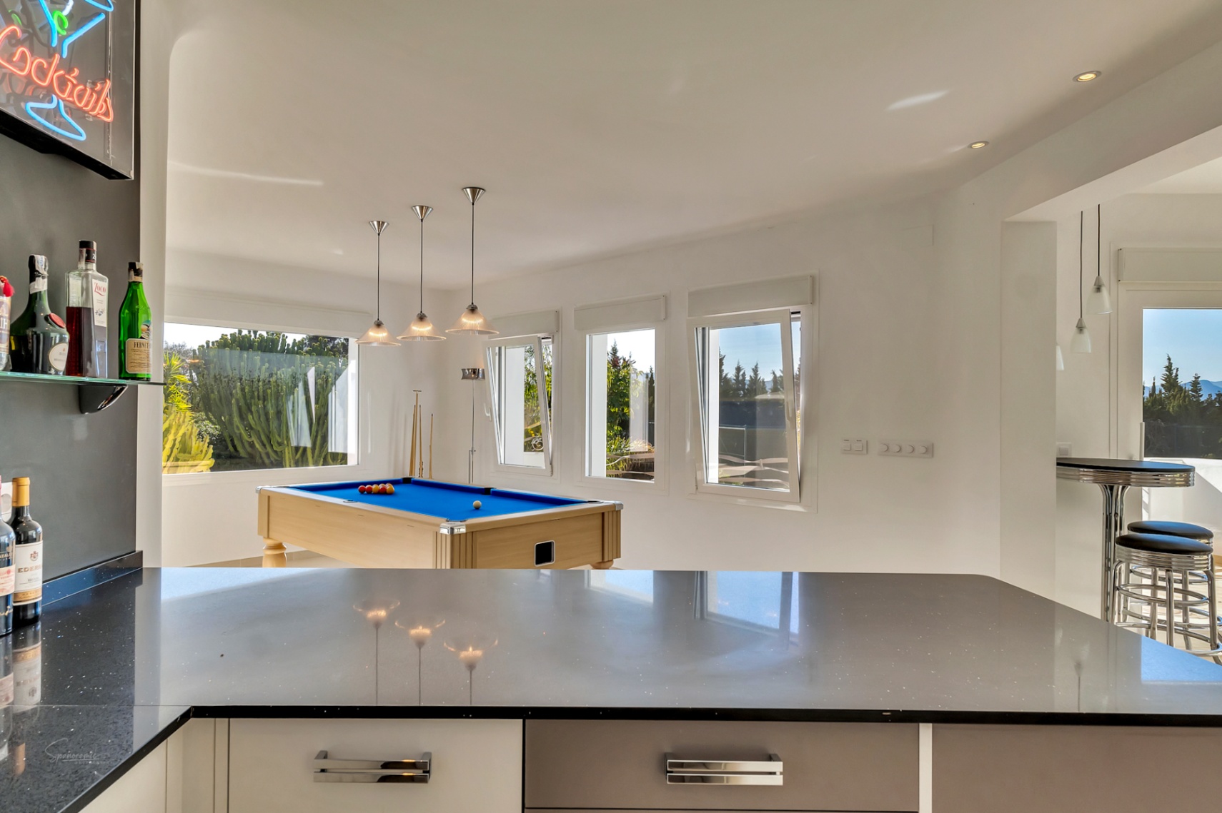 Sala de ocio luminosa con mesa de billar, zona de bar integrada con encimera oscura y amplios ventanales que conectan con el exterior.