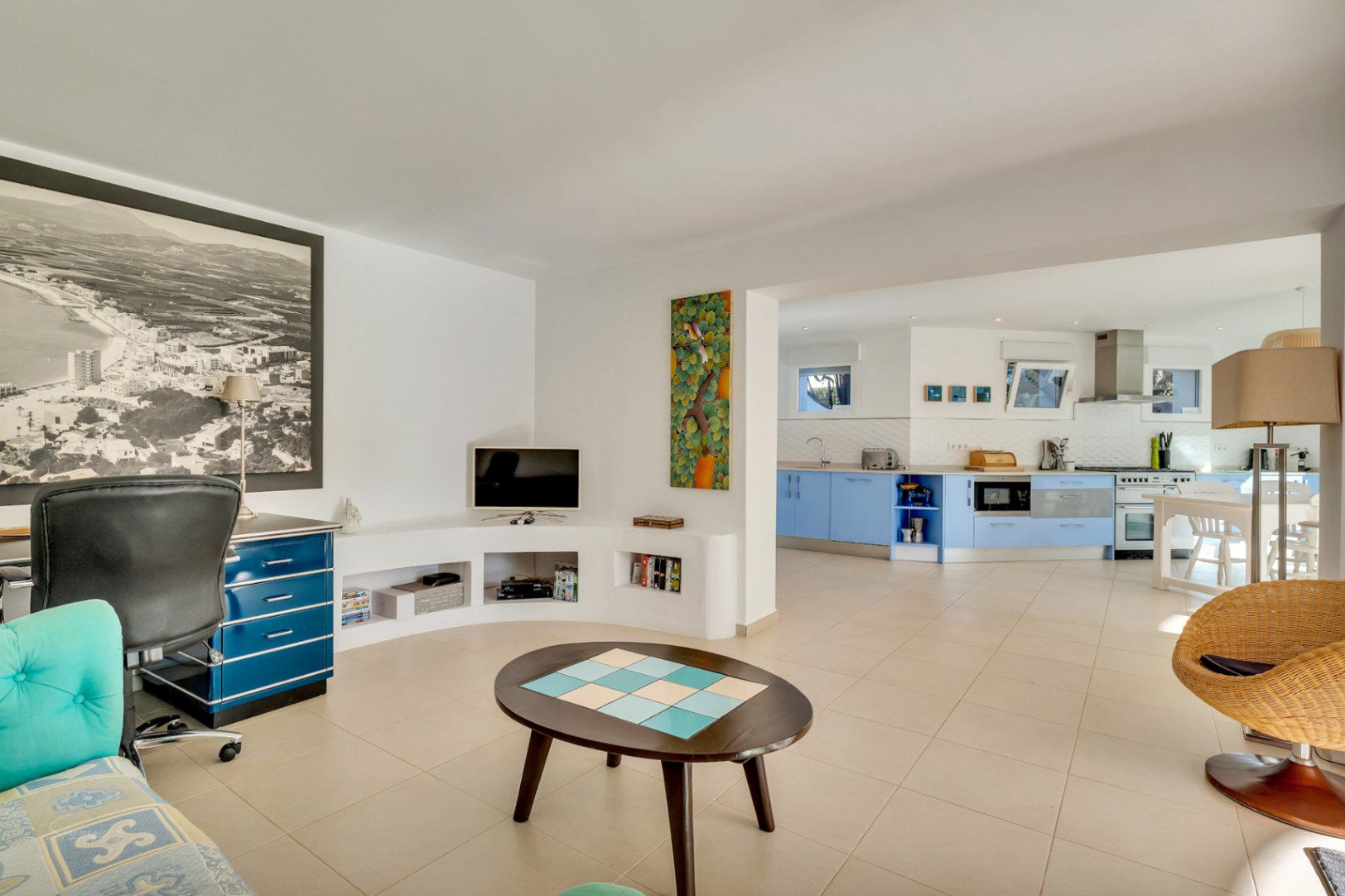 Salón diáfano con zona de despacho, estantería curva integrada y acceso a una cocina moderna con armarios azules y electrodomésticos de acero inoxidable.