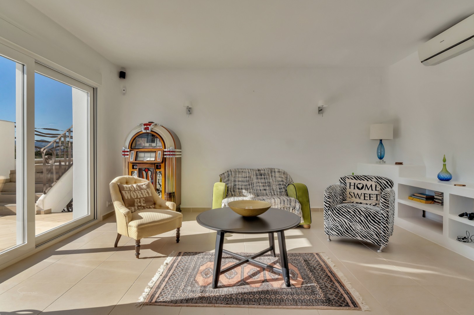 Salón luminoso con mobiliario ecléctico, una gramola clásica y estanterías integradas, conectado a la terraza exterior a través de amplios ventanales correderos.